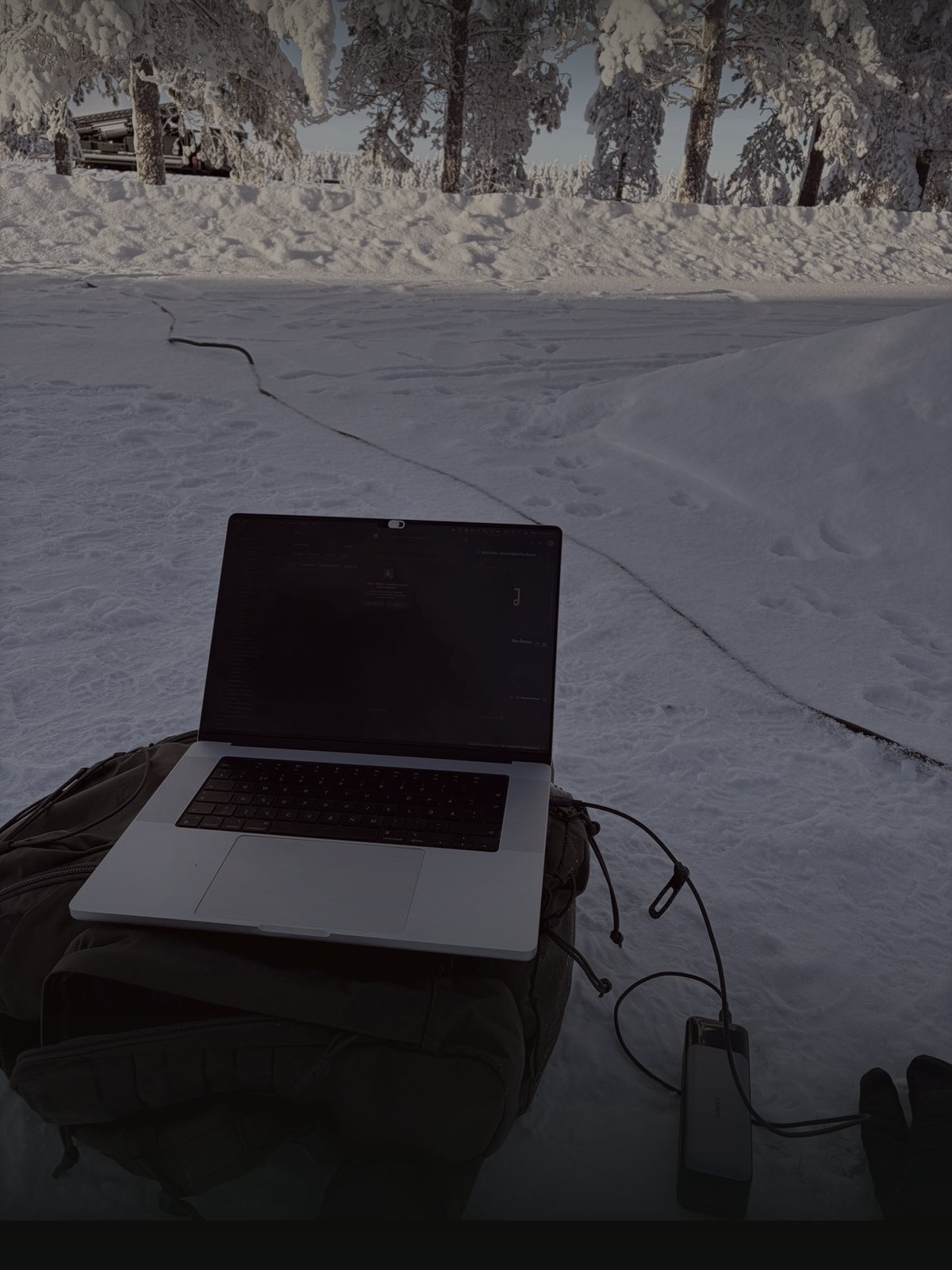 Laptop on a field pack in deep snow, power bank and cable running across the ground, treeline behind